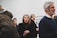 A group of people gathered in what appears to be an art gallery or exhibition space. Several individuals are visible, dressed in winter clothing. A woman with long hair stands prominently in the foreground, looking engaged. In the background, there are white walls and framed artworks, indicating a formal setting.