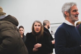 A group of people gathered in what appears to be an art gallery or exhibition space. Several individuals are visible, dressed in winter clothing. A woman with long hair stands prominently in the foreground, looking engaged. In the background, there are white walls and framed artworks, indicating a formal setting.