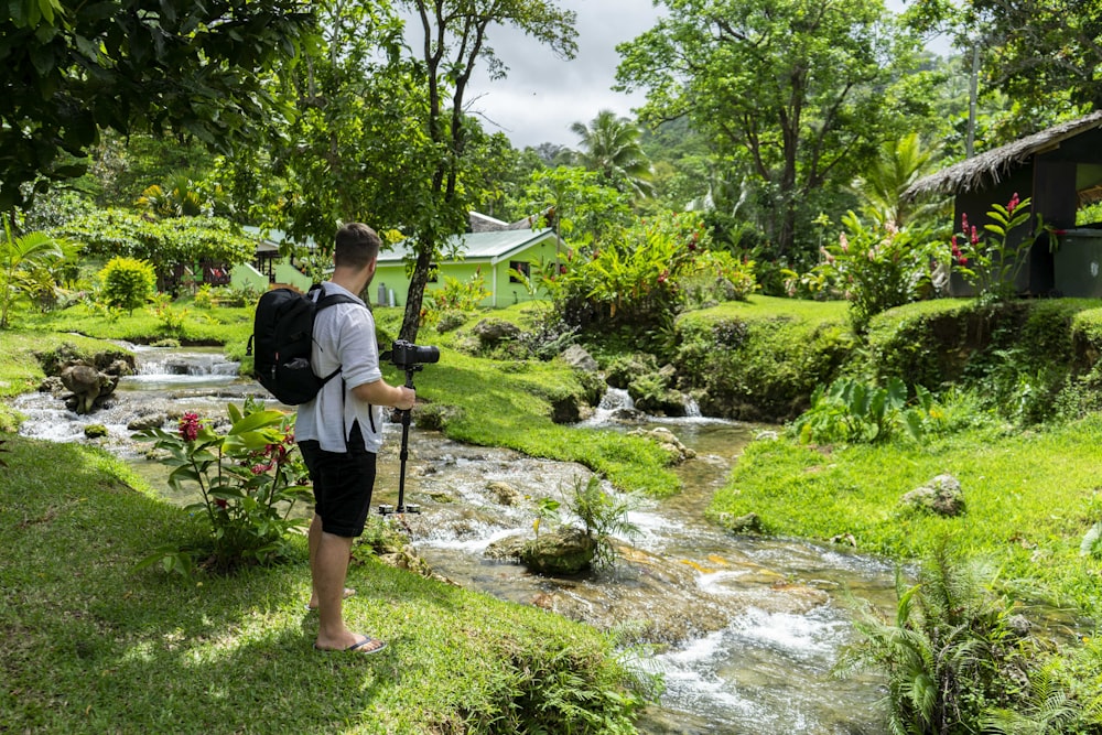 man in grey shirt and black shorts standing by the river bank near house during daytime