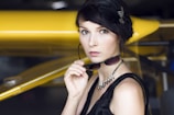A woman with dark hair styled elegantly and adorned with a feathered accessory holds a pair of sunglasses. She wears a black dress and a stylish necklace, standing in front of a yellow airplane wing.