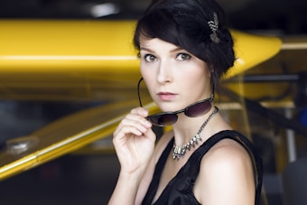 A woman with dark hair styled elegantly and adorned with a feathered accessory holds a pair of sunglasses. She wears a black dress and a stylish necklace, standing in front of a yellow airplane wing.