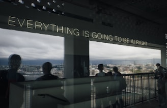 Lighted text on a window reads 'Everything is going to be alright' with a reflective view of a cityscape and cloudy sky. Silhouettes of people appear in the foreground.