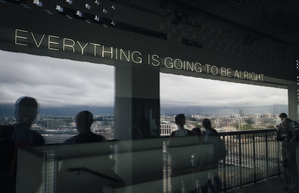 Lighted text on a window reads 'Everything is going to be alright' with a reflective view of a cityscape and cloudy sky. Silhouettes of people appear in the foreground.