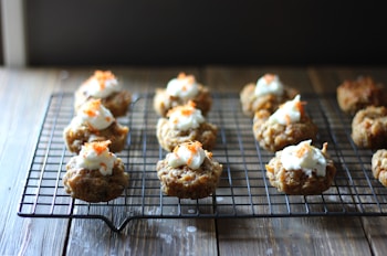 A collection of small baked goods topped with cream and orange zest is arranged in rows on a cooling rack. The baked items appear golden-brown and are placed on a dark wire rack over a wooden surface.