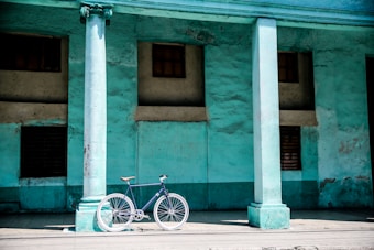 A vintage bicycle with a leather saddle is leaning against a weathered teal-colored wall, framed by two large classical columns. The wall has a textured, aged appearance, with small windows and rusty metal details, conveying a sense of history and nostalgia.