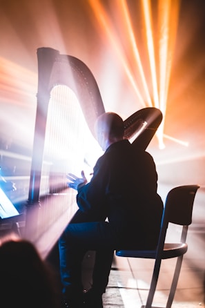 A person is playing the harp, illuminated by dramatic and colorful stage lighting. Beams of light create a vibrant, ethereal atmosphere, casting shadows and highlighting the musician and instrument.