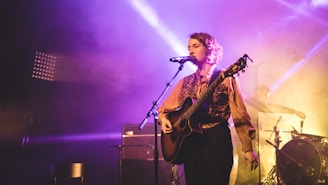A musician with curly hair is playing an acoustic guitar and singing into a microphone on stage. The scene is lit with vibrant purple and yellow stage lights creating a dramatic atmosphere. An additional band member is visible in the background behind a drum set.