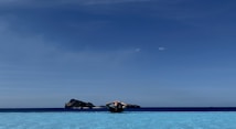 A person is lying back on an inflatable pool float in a serene infinity pool. The calm water merges seamlessly with the horizon, beyond which lies a tranquil sea and distant rocky islands. The sky is clear with shades of deep blue.