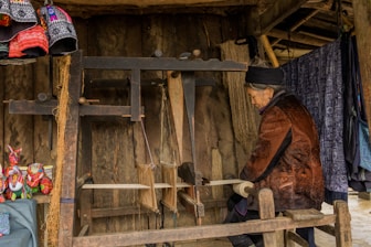An elderly person is sitting and working on a traditional wooden loom in an indoor setting with wooden walls. Colorful textiles and fabrics adorn the area, with some hanging and folded nearby. Vibrant, patterned clothing items and small, colorful stuffed animals are displayed to the left. A woven basket and some draped fabric create a warm, artisanal atmosphere.