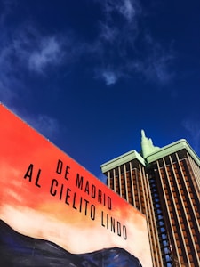 A billboard with the text 'De Madrid al Cielito Lindo' is set against a richly colored sky. The scene includes a building with a modern design, featuring a green and brown color scheme.