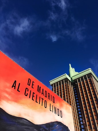 A billboard with the text 'De Madrid al Cielito Lindo' is set against a richly colored sky. The scene includes a building with a modern design, featuring a green and brown color scheme.