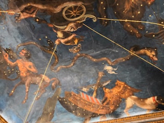 A richly detailed ceiling mural depicting mythological scenes featuring celestial figures, mythical creatures, and a ship. The artwork includes a centaur holding a bow, a chariot with a figure in armor, and a large snake amidst a starry backdrop. The imagery combines classical mythology with astrological elements, creating a grand and immersive visual experience.