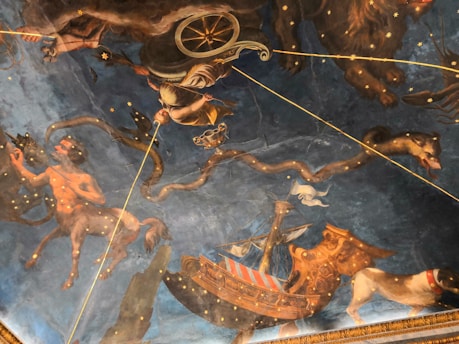 A richly detailed ceiling mural depicting mythological scenes featuring celestial figures, mythical creatures, and a ship. The artwork includes a centaur holding a bow, a chariot with a figure in armor, and a large snake amidst a starry backdrop. The imagery combines classical mythology with astrological elements, creating a grand and immersive visual experience.