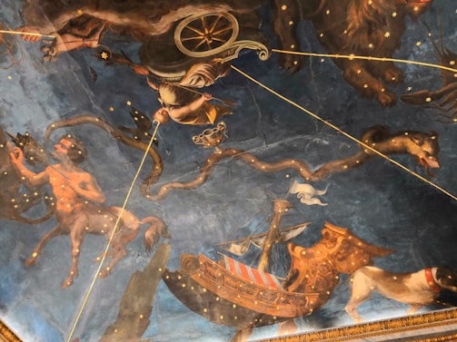 A richly detailed ceiling mural depicting mythological scenes featuring celestial figures, mythical creatures, and a ship. The artwork includes a centaur holding a bow, a chariot with a figure in armor, and a large snake amidst a starry backdrop. The imagery combines classical mythology with astrological elements, creating a grand and immersive visual experience.