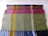 Three woven textiles stacked on top of each other, each featuring a distinct pattern and color scheme. The uppermost fabric has a checkered design with orange, blue, and yellow hues. The middle piece has a similar checkered pattern in shades of pink. The bottom textile displays a green and blue speckled pattern with long fringes at the end.