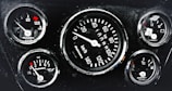 A cluster of five automotive gauges arranged in a dashboard. Central gauge displays speed in kilometers per hour, surrounded by smaller gauges indicating temperature, oil pressure, fuel level, and battery status. Each gauge features red indicators against a black background, with white numerals and markings.