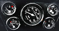 A cluster of five automotive gauges arranged in a dashboard. Central gauge displays speed in kilometers per hour, surrounded by smaller gauges indicating temperature, oil pressure, fuel level, and battery status. Each gauge features red indicators against a black background, with white numerals and markings.