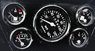 A cluster of five automotive gauges arranged in a dashboard. Central gauge displays speed in kilometers per hour, surrounded by smaller gauges indicating temperature, oil pressure, fuel level, and battery status. Each gauge features red indicators against a black background, with white numerals and markings.