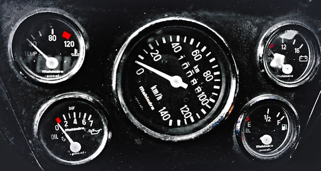 A cluster of five automotive gauges arranged in a dashboard. Central gauge displays speed in kilometers per hour, surrounded by smaller gauges indicating temperature, oil pressure, fuel level, and battery status. Each gauge features red indicators against a black background, with white numerals and markings.