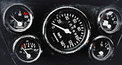 A cluster of five automotive gauges arranged in a dashboard. Central gauge displays speed in kilometers per hour, surrounded by smaller gauges indicating temperature, oil pressure, fuel level, and battery status. Each gauge features red indicators against a black background, with white numerals and markings.