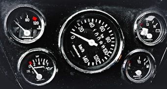 A cluster of five automotive gauges arranged in a dashboard. Central gauge displays speed in kilometers per hour, surrounded by smaller gauges indicating temperature, oil pressure, fuel level, and battery status. Each gauge features red indicators against a black background, with white numerals and markings.