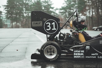 A driver wearing a helmet is seated in a small racing car with the number 31 marked on the rear wing. The car is parked on a wet pavement near a forested area, with trees visible in the background. The vehicle has logos from various sponsors including SKF, Lucas Oil, and Hoosier Tire.