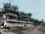 A rustic coastal structure with a thatched roof and wooden balcony sits amidst tall palm trees. People are visible on the balcony and surrounding area. A small cart filled with colorful goods is parked on a rocky surface nearby.