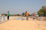 A group of people is gathered around a sand pit on an outdoor athletics track, likely engaging in or observing long jump practice. One individual appears to be mid-jump while others are standing nearby, possibly as coaches or spectators.