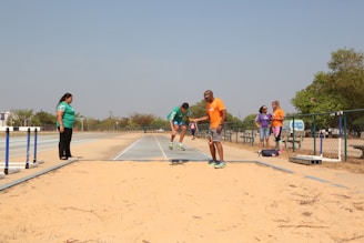 A group of people is gathered around a sand pit on an outdoor athletics track, likely engaging in or observing long jump practice. One individual appears to be mid-jump while others are standing nearby, possibly as coaches or spectators.