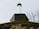 A close-up view of a chimney on top of a building with a moss-covered roof. The sky is overcast, giving a sense of an early spring or late autumn day. In the background, leafless branches of a tree can be seen.