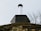 A close-up view of a chimney on top of a building with a moss-covered roof. The sky is overcast, giving a sense of an early spring or late autumn day. In the background, leafless branches of a tree can be seen.