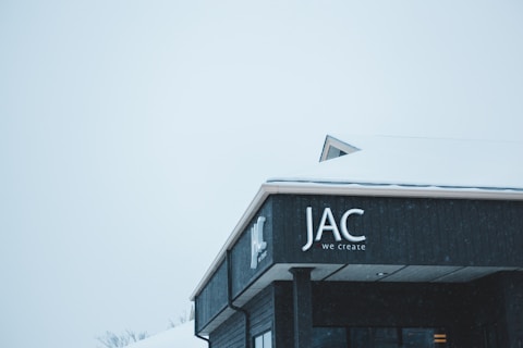 A dark-colored building with a sign that reads 'JAC we create' is partially covered in snow. The roof is white with a layer of snow, and the sky appears overcast with a soft, light gray tone.