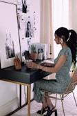 A woman is seated at a stylish, modern desk, adorned with various items including a small brown handbag, a plant, and some magazines featuring fashion and lifestyle images. The wall behind the desk is decorated with black and white photographs and inspirational images. She is wearing a green dress with white patterns and dark heeled shoes, and she appears to be working or studying.