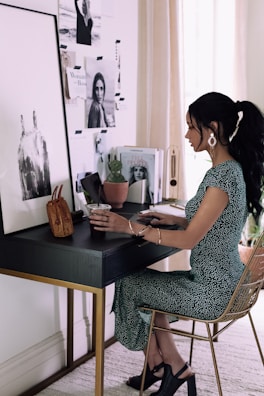 A woman is seated at a stylish, modern desk, adorned with various items including a small brown handbag, a plant, and some magazines featuring fashion and lifestyle images. The wall behind the desk is decorated with black and white photographs and inspirational images. She is wearing a green dress with white patterns and dark heeled shoes, and she appears to be working or studying.