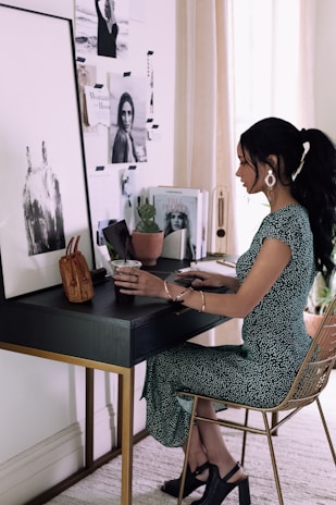 A woman is seated at a stylish, modern desk, adorned with various items including a small brown handbag, a plant, and some magazines featuring fashion and lifestyle images. The wall behind the desk is decorated with black and white photographs and inspirational images. She is wearing a green dress with white patterns and dark heeled shoes, and she appears to be working or studying.