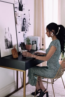 A woman is seated at a stylish, modern desk, adorned with various items including a small brown handbag, a plant, and some magazines featuring fashion and lifestyle images. The wall behind the desk is decorated with black and white photographs and inspirational images. She is wearing a green dress with white patterns and dark heeled shoes, and she appears to be working or studying.