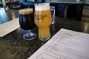 A dark and a light beer glass are placed on a table in a bar setting. The glasses are positioned next to a menu that lists various pizza options. The background features a bar counter with a glass of water.