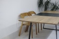 A minimalist setting with a wooden table and chairs against a white wall. The table has a modern design with black legs and the chairs match in color and style. In the background, there are green potted plants that add a touch of nature to the space.