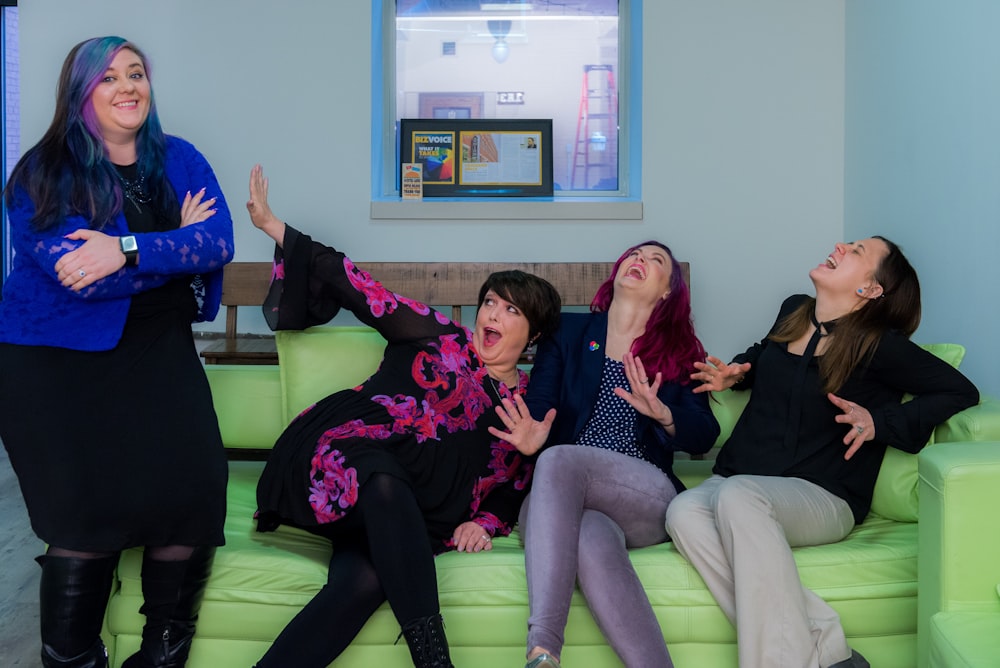 three women laughing while sitting on green sofa beside woman standing