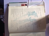 An open recipe book lies on a white surface, revealing a slightly worn page with a detailed contents list. The contents are categorized into different sections like 'ensaladas' and 'platos principales,' with page numbers for various recipes. A shadow is cast over part of the page, and a partially visible white cable is draped across the top. Another book is visible beneath, with colorful images peeking out.