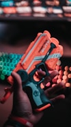A hand holding a brightly colored toy blaster with the words 'FIRESTRIKE ELITE' visible on its side. The background features blurry, colorful lights resembling a gaming setup or a computer keyboard.