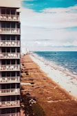 A tall building with multiple balconies is seen on the left side, adjacent to a sandy beach that stretches along the coastline. The ocean is visible to the right, with gentle waves lapping the shore. People are scattered along the beach, enjoying the sun and sea. The sky is partly cloudy with a vibrant blue tone.