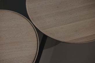 Two round wooden tabletops with a light wood grain texture, positioned closely together over a dark background. The grain patterns are subtly visible, emphasizing the natural qualities of the wood.