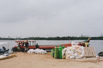 A dockside scene features a wooden boat moored by the shore, surrounded by people engaged in loading or unloading activities. Numerous white sacks are piled nearby on the sandy ground, along with green gas cylinders. The backdrop includes a body of water and a distant view of industrial structures against a cloudy sky.