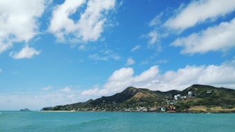 A scenic coastal landscape featuring a turquoise sea and a mountainous coastline under a partly cloudy blue sky. Small buildings are nestled along the shore, surrounded by lush green vegetation.