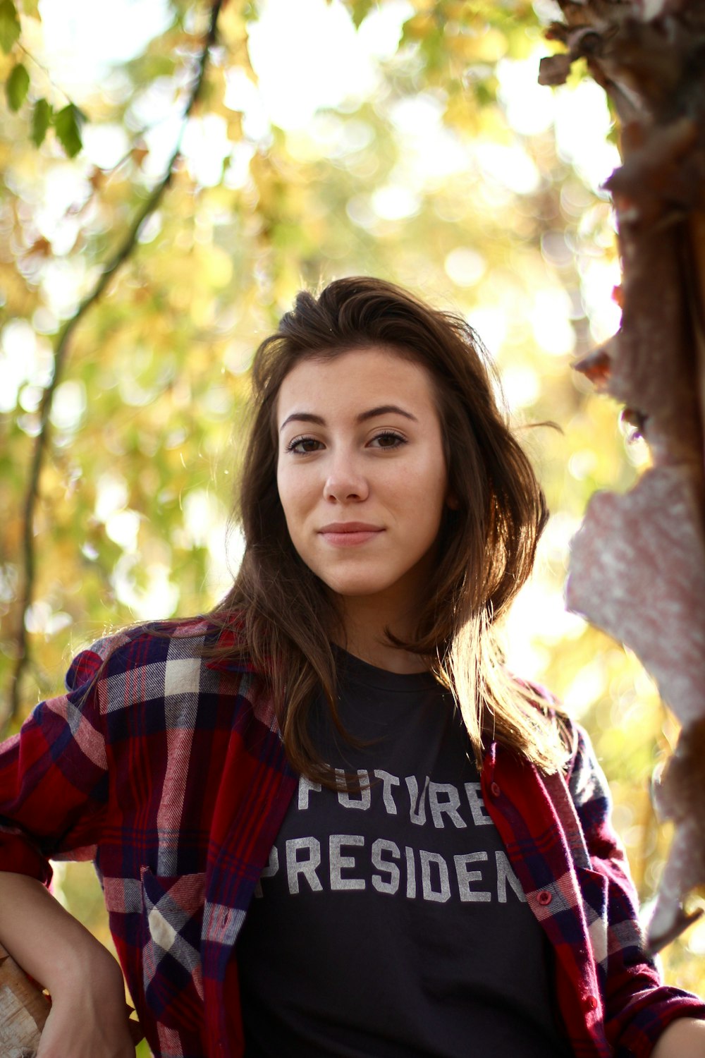 woman wearing black shirt and red-white-blue plaid jacket across blurry tree background