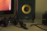 A close-up view of a gaming setup includes a black video game controller with a connecting cable, positioned next to a speaker with a yellow center. A part of a television screen displaying a person wearing an orange shirt is visible. The overall setting is on a light-colored tabletop, and there are additional electronic accessories partially visible.