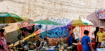 A lively street market scene with vendors selling plants and goods under colorful umbrellas. The background features a wall adorned with vibrant graffiti art depicting a stylized animal character in bright colors. Several people are walking and engaging with the market stalls, creating a bustling and dynamic atmosphere.