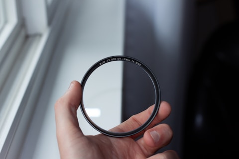 A hand is holding a circular camera filter near a window. The filter is labeled with markings that include 'B+W' and 'UV-Haze'. The background shows an out-of-focus window and wall, creating a soft lighting effect.