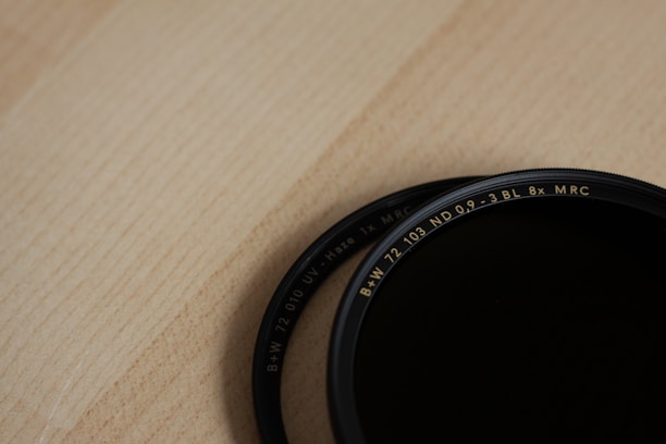 A close-up view of two circular camera lens filters placed on a wooden surface. The filters have detailed text inscribed on the outer rims indicating their specifications.